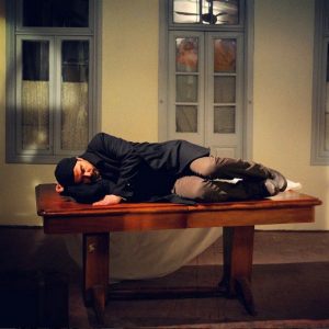 علی مصفا خوابیده روی یک میز در صحنه ای از فیلم در دنیای تو ساعت چند است